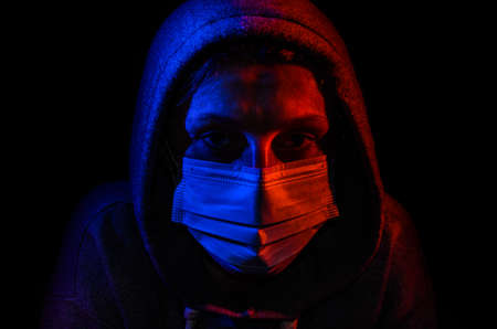 Portrait of a young woman in a hood, wearing a protective medical mask, illuminated by red and blue light, isolated on black backgroundの写真素材