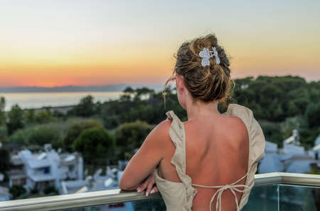 Young charming woman in a dress with a bare back stands on the balcony overlooking the sea at the sunset timeの写真素材