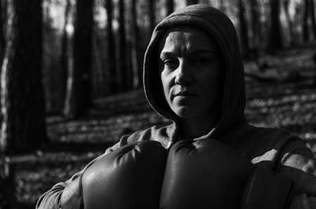 Young beautiful woman boxer in a tracksuit with a hood on his head in red boxing gloves trains in the forest in natureの写真素材