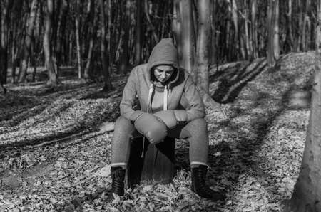 Young charming girl, in a tracksuit with a hood on her head, in red boxing gloves, is resting after training in women's boxing in the autumn forest in natureの写真素材