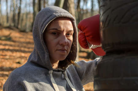Young beautiful woman boxer in a tracksuit with a hood on his head in red boxing gloves trains in...