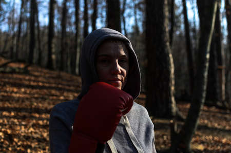 Young beautiful woman boxer in a tracksuit with a hood on his head in red boxing gloves trains in...