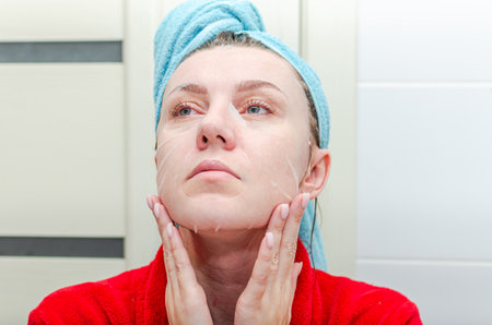Woman doing herself a spa beauty treatment by laying down a moisturizing sheet mask in the bathroom, after a showerの写真素材