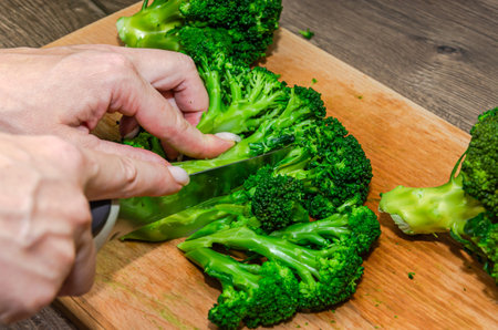 Female hands cutting fresh green broccoli on a wooden board with a knifeの写真素材