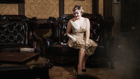 a girl in a gold dress with a diadem on her head, sits on a leather armchair, next to a gun and...