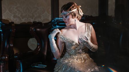 a girl in a gold dress with a diadem on her head, holds a glass and sits on a leather chair, with...