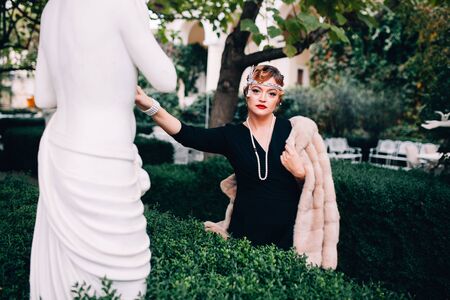 a gorgeous woman in a fur cape and a diadem on her head walks through her garden surrounded by Greek statues, touches the statue with her handの写真素材