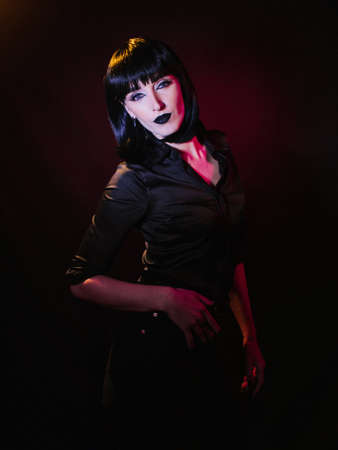 A woman in a black blouse, on a black background with colored light, looks curiously at the camera