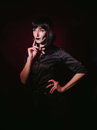 A girl with black hair, dark makeup and black clothes posing with her hands in the frame. Her gaze...