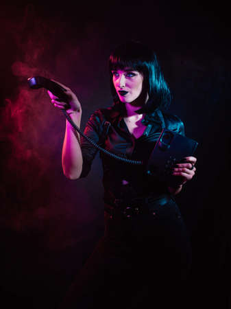 a girl in colored light, with black hair in a harness, points to the side with a receiver from a landline phoneの写真素材