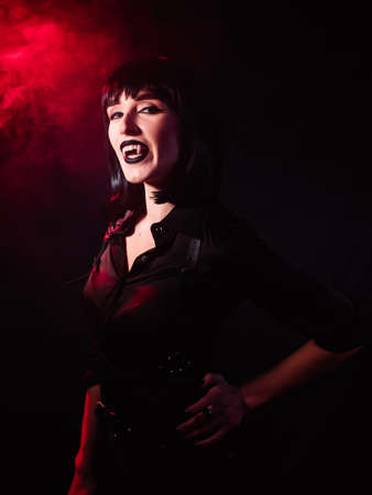 Girl on a black background with red light and artificial smoke in the image of a vampire. She looks haughtily and laughs with her mouth open, showing her fangs.の写真素材