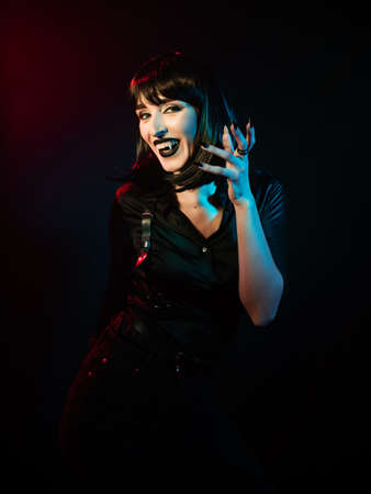 a girl with vampire fangs with black hair in a black shirt and harness raised her hand with long...