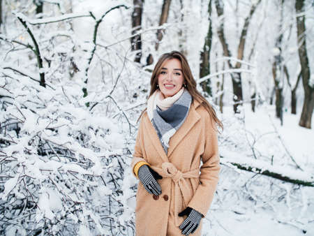 a girl in a beige coat with a scarf and gloves stands among the icy branches in winter in the parkの写真素材