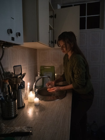 a young woman washes dishes in a kitchen without electricity in a sweater, and with a flashlight on her head by candlelight.の写真素材