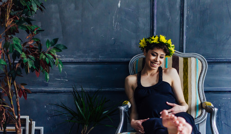 Pregnant woman sits in an armchair by the window next to tree. She looks thoughtfully at her bellyの写真素材