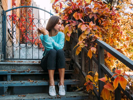 Beautiful young girl in a blue sweater, sitting on the steps of the veranda, enjoying the autumn landscape.の写真素材