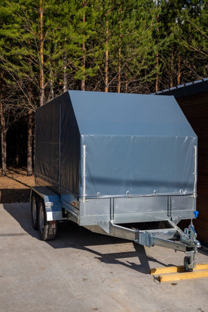 Grey covered utility cargo trailer parked on a driveway near a pine forest. Concept: transport logistics