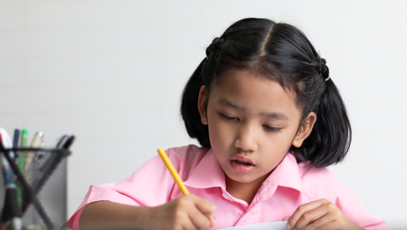 Close up the little girl is doing homework intently. Children use a yellow pencil is writing a...