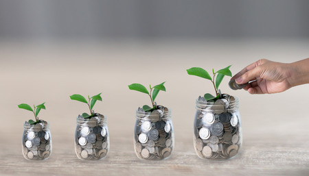Hand putting a coin in a jar which a growing plant in coins on wooden table with blurred backgroud and copy spaceの写真素材