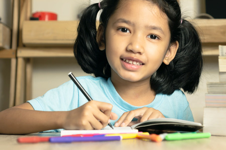 The little asian girl is writing a book on the wooden table with smile and happinessの写真素材