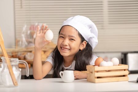 A little Asian girl wearing a white chef at is staring the duck egg in holding her hand and smiling on a white cooking table in the kitchen. Select focus shallow depth of field.の写真素材