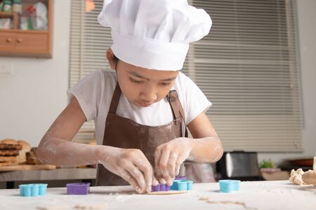 The little asian girl wore a white chef hat and a brown apron. The kid making cookies in the...