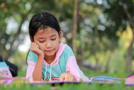 The portrait Asian little girl reading a book in the garden. The kid takes a rest outside on the grass with sunray on the natural. The environment in the park for relaxation.の写真素材