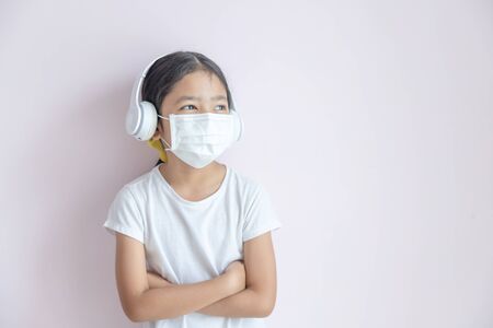 A little Asian girl wearing a medical protective mask and earphones. The child girl standing cross one's arm on the color background. The epidemic of the flu, Coronavirus or COVID-19 and illness concept.の写真素材