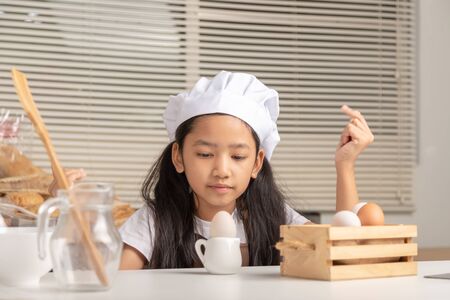 A little Asian girl wearing a white chef hat is staring at a duck egg on a small jug and puts on a white cooking table in the kitchen.の写真素材
