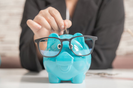 Close-up a blue piggy bank wearing black glasses on the wooden table. The symbol of saving money and wealth financial. A businesswoman hand putting coin into a piggy bank.の写真素材