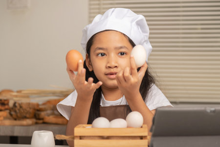 A little Asian girl wearing a white chef hat is choosing between a duck egg and a chicken egg to make a bakery on a white cooking table in the kitchen.の写真素材