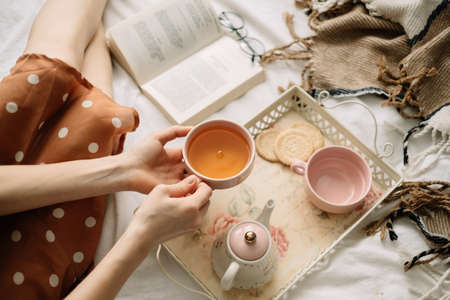 A woman holds a cup of hot tea on the bed at home and reads a book, home leisure in coziness and comfort.の写真素材
