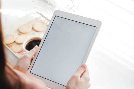 Woman holding the tablet with a white screen mock up, and drinking coffee at home on a sunny day. High quality photoの写真素材