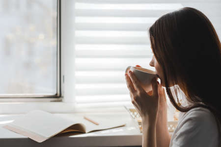 Woman drinking tea sitting by the window and takes notes on a sunny spring morning. High quality...