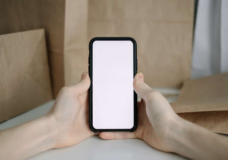 Woman holds smartphone with white blank screen, packages with purchases on the table. Online shopping concept. High quality photoの写真素材