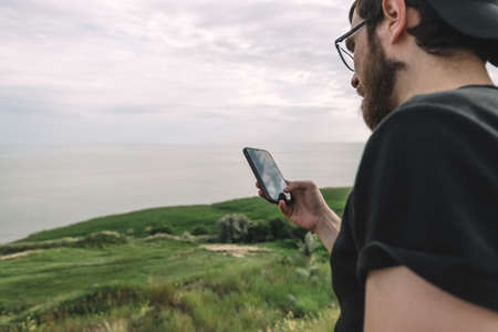 Young bearded caucasian man uses a smartphone on the nature near the sea on a sunny day. High...