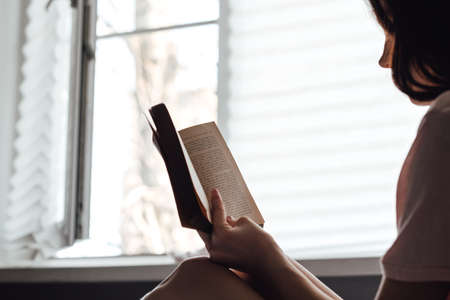 Woman reads a book at home near the window.