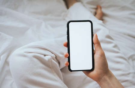 Woman holding a smartphone with a white screen, sitting on the bed at home on a sunny day. Mock up.の写真素材
