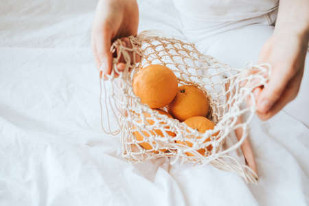 Woman takes oranges from a net eco bag on a white bed at home.の写真素材