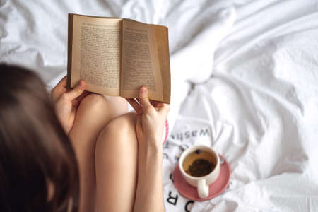 Woman sitting on a white bed at home and reading book and drink tea.の写真素材