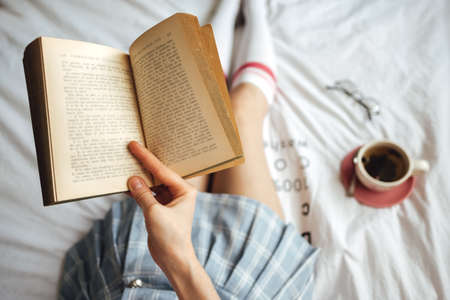 Young caucasian woman sitting on a white bed at home reading a book and drink tea. High quality photoの写真素材