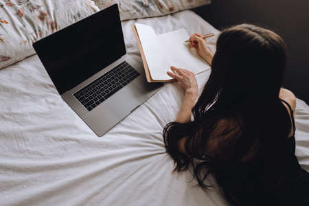 Young woman lies on bed with laptop and notebook and takes notes. Distance education, remote work.の写真素材