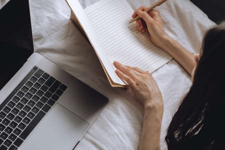 Young woman lies on bed with laptop and notebook and takes notes. Distance education, remote work.の写真素材