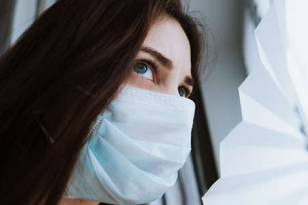 Young female in a medical protective mask looks out the window.Coronavirus Quarantine Concept.の写真素材