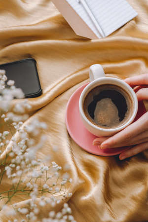 The cup of coffee in hands on a golden satin fabric with gypsophila flowers. Spring morning.の写真素材