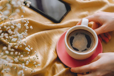 The cup of coffee in hands on a golden satin fabric with gypsophila flowers. Spring morning. Phone on the background.の写真素材