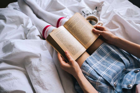 Young caucasian woman sitting on a white bed at home reading a book and drink tea. High quality photoの写真素材