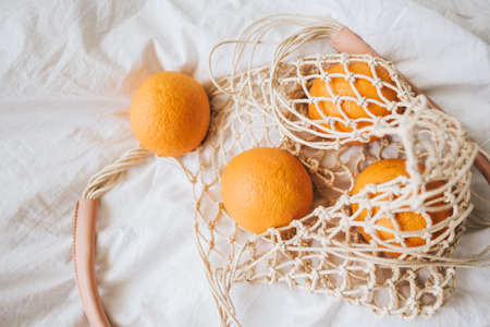 Mesh eco bag with fresh oranges on a white cloth at home. Top view.