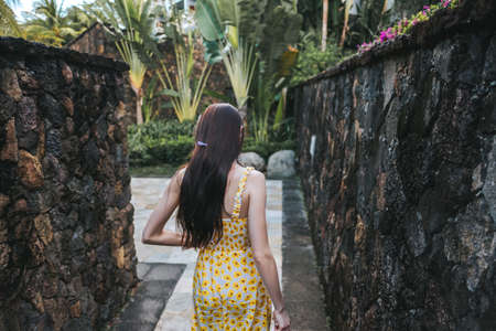 Young beautiful brunette woman in a yellow dress walks along a stone path in a tropical park, around a palm tree. outdoor recreation in summer. High quality photoの写真素材