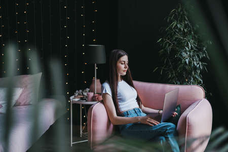 Beautiful young caucasian woman working with laptop computer, sitting on a pink sofa at home near...